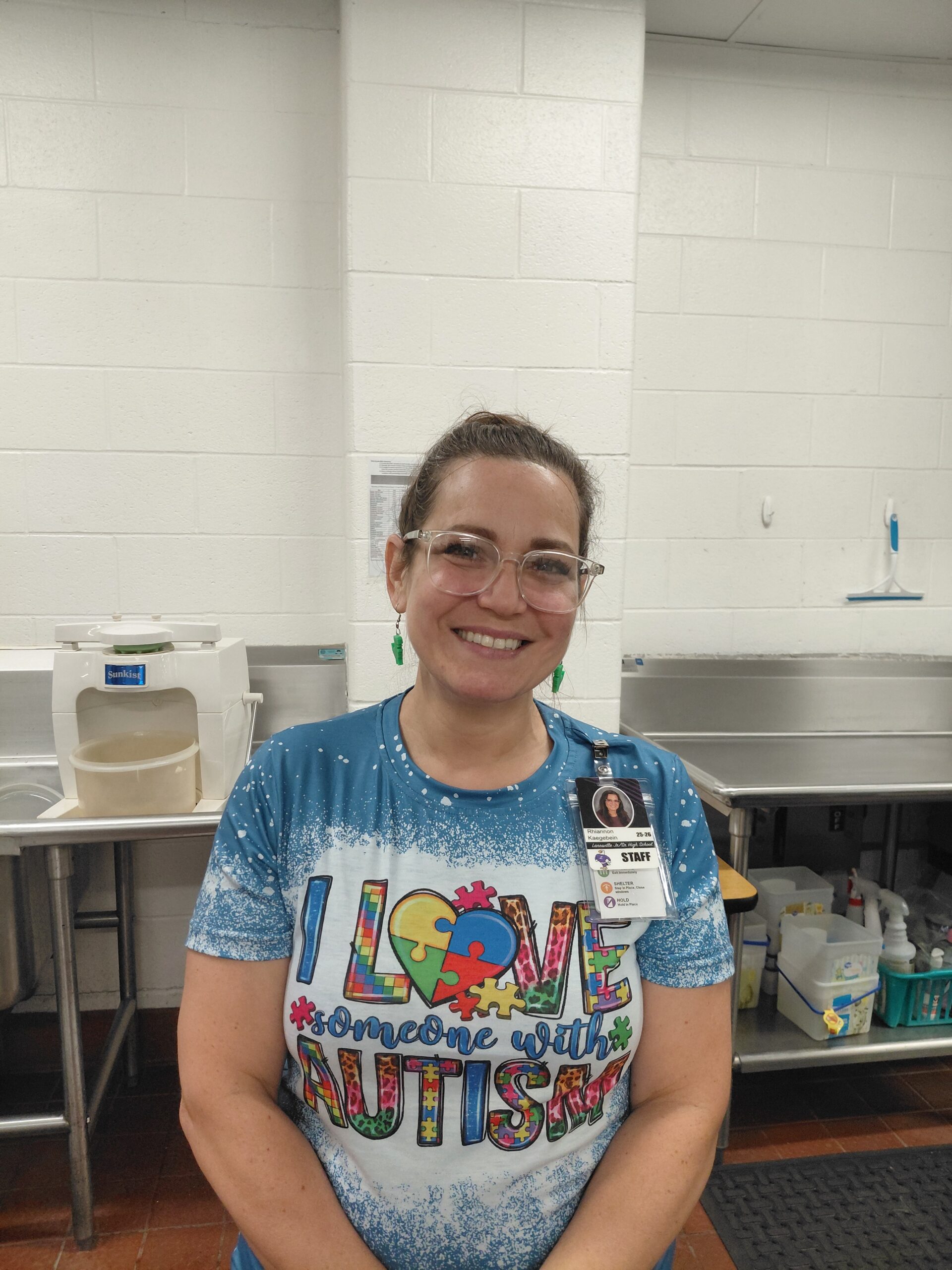 Smiling female staff member in a kitchen, wearing safety glasses and a name badge, with a colorful shirt that says 'Love someone with autism'.