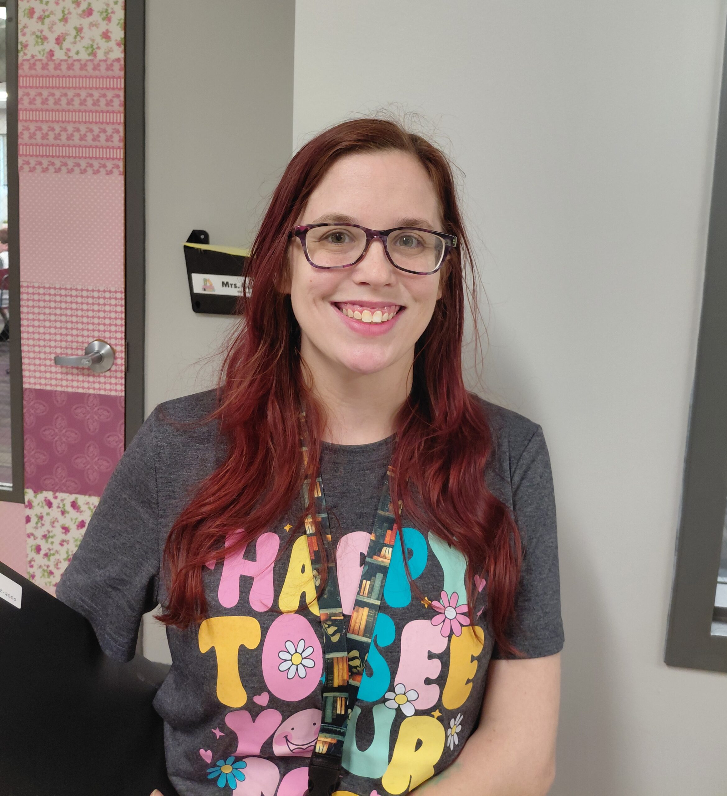 Smiling woman with red hair and glasses wearing a gray graphic tee indoors, pink patterned wall behind her.
